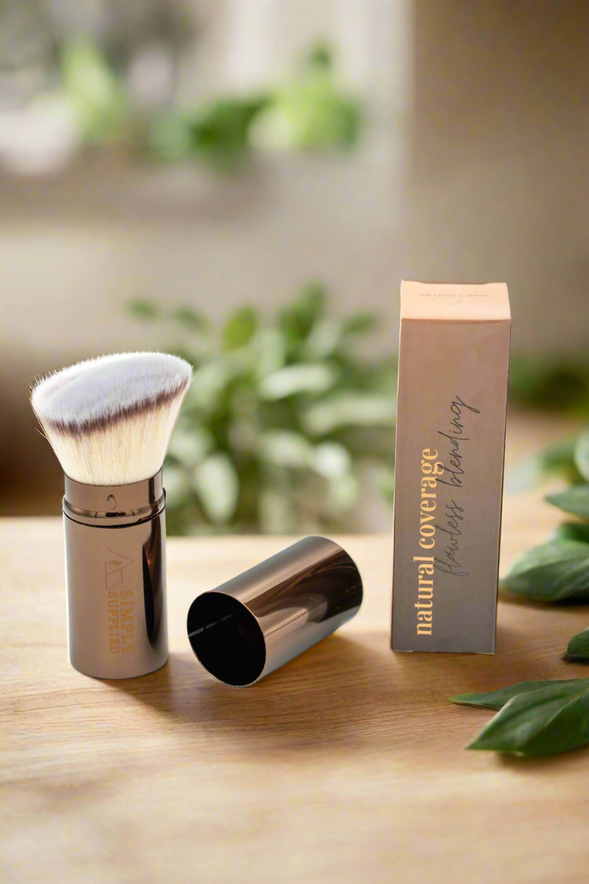 Makeup brush with brown handle and white bristle head, next to its packaging with a planty green background