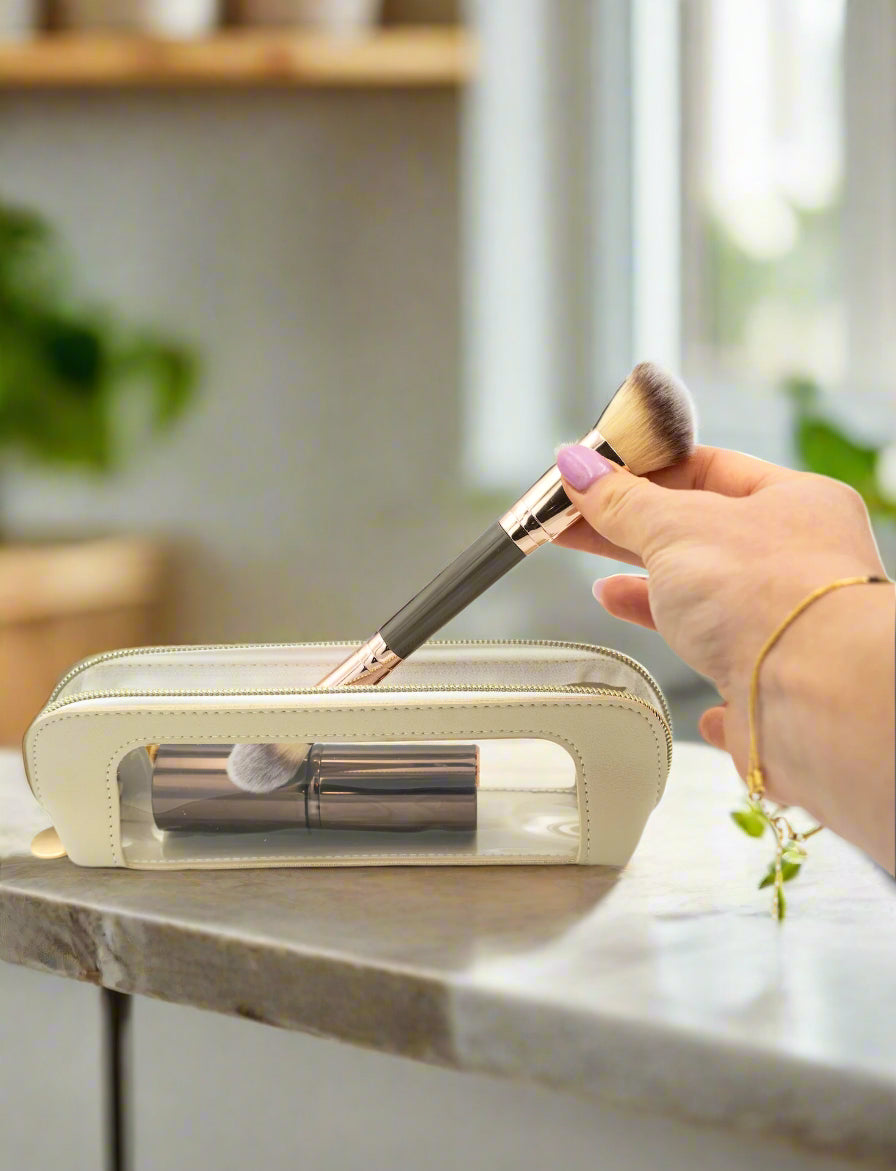 Hand holding a makeup brush over a transparent makeup case on a light gray background